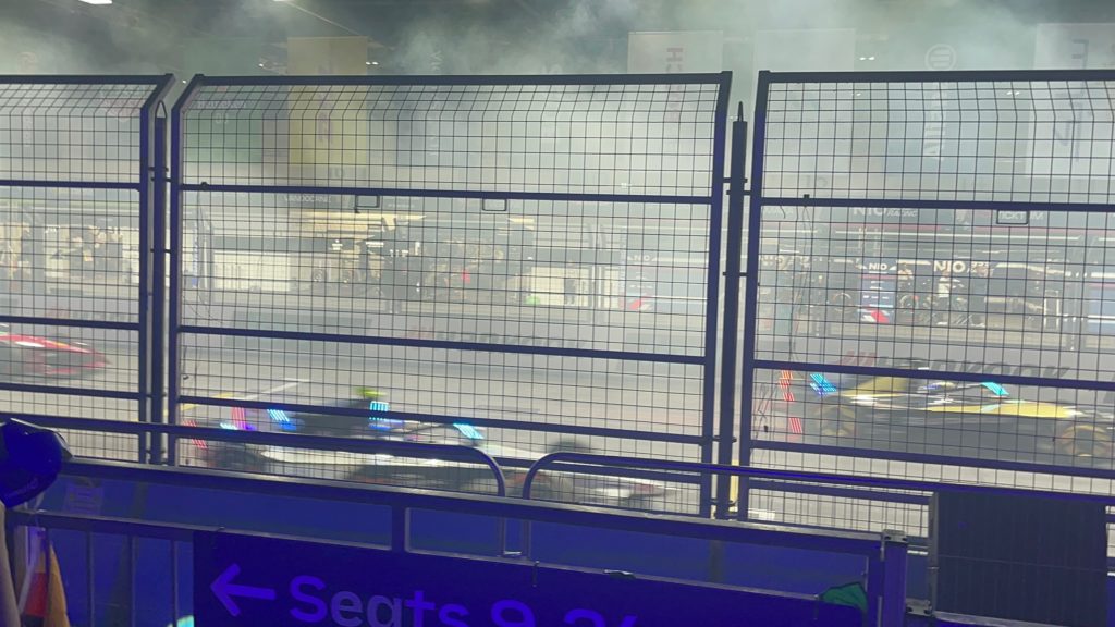 Photograph of the Formula e grid, Saturday 29th July 2023, taken from grandstand 5, behind the barriers. There is smoke on the grid. The team pits are faintly visible.