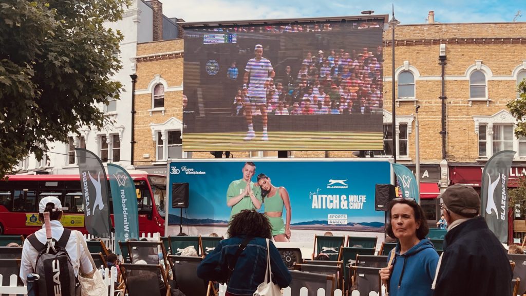 A big television screen in Wimbledon town centre showing The Championships. There are deckchairs in front of the screen with a few people sat on them watching.