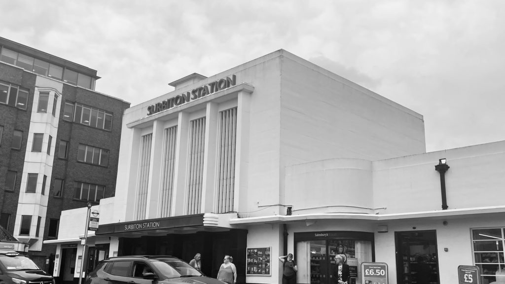 View of the front of Surbiton staton with a taxi and car in front of the station buildings. A few people waiting. Picture in black and white.