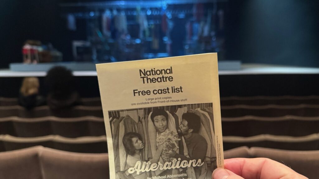 This image shows a cast list sheet for a play called "Alterations" by Michael Abbensetts at the National Theatre in London. Someone is holding the beige/cream-coloured programme or cast list in front of theatre seating. The cover features a black and white photograph showing three people among clothing on hangers in what looks like a clothing shop. The background of the image shows rows of theatre seats and a dimly lit stage area or set in the distance, indicating this was taken from an audience perspective before or after the performance.