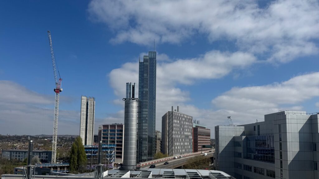 This image shows the modern skyline of White City in London. The view captures several contemporary high-rise and mid-rise buildings under a bright blue sky with scattered white clouds. A construction crane is visible on the left side, indicating ongoing development in the area. The skyline features a mix of architectural styles, including a prominent glass skyscraper in the centre, cylindrical structures, and angular office buildings with glass facades.