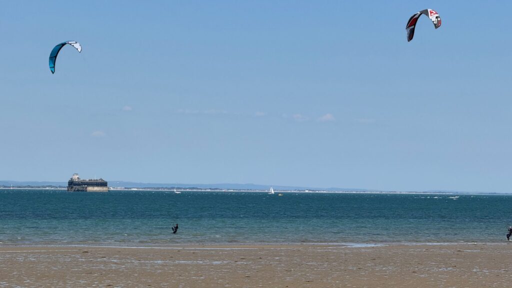 This image shows a coastal scene at Ryde on the Isle of Wight, featuring a sandy beach with calm blue waters under a partly cloudy sky. Two colourful kites - one teal and one with red, white and blue stripes - are flying above the water, suggesting kitesurfing activity. Several small sailing boats can be seen dotting the horizon, whilst the foreground shows the sandy shoreline. The scene captures a typical day of water sports and leisure activities along this popular stretch of the Solent.