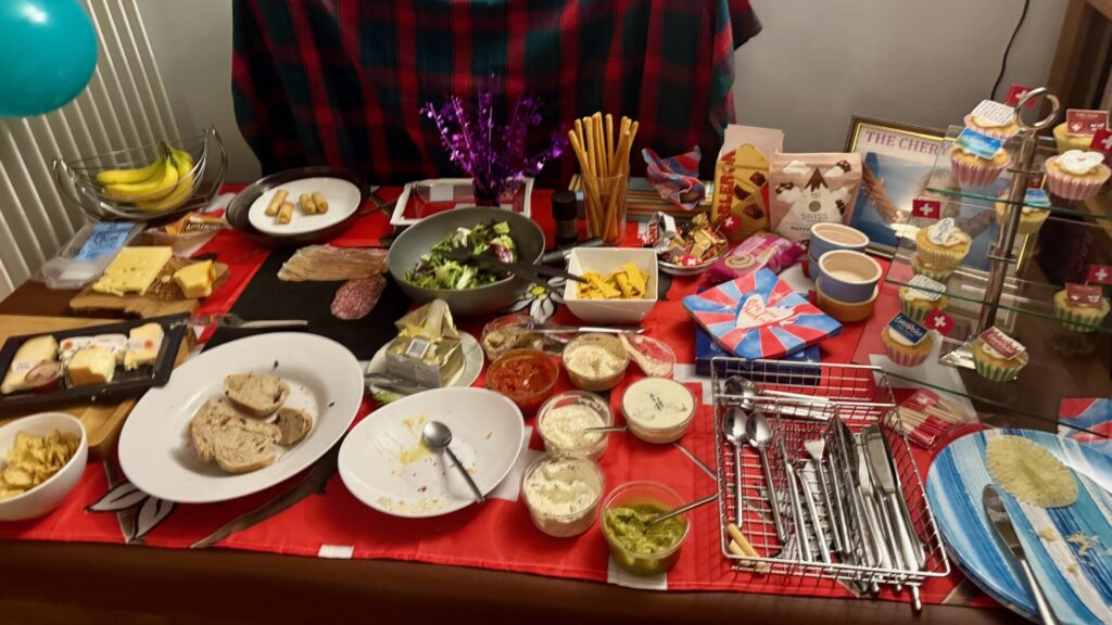 This image shows a festive Eurovision party buffet spread across a dining table covered with red and blue checkered tablecloths. The table is laden with various foods including cheese and charcuterie boards, salads, dips, bread, and Swiss-themed items (with small Swiss flags visible). There are also cupcakes on a tiered stand, wrapped presents, purple decorative elements, and party accessories scattered throughout. The abundant spread suggests a well-prepared celebration with both savoury and sweet options for guests.