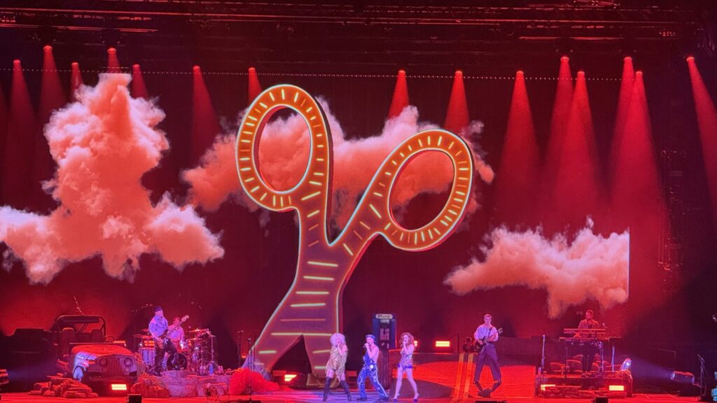 This vibrant concert photograph captures the Scissor Sisters performing at The O2 in London on 23rd May, featuring a spectacular stage design dominated by an enormous illuminated scissor prop that towers above the performers. The stage is bathed in dramatic red lighting with atmospheric smoke effects, creating a theatrical backdrop of concert lighting rigs and towering equipment. Band members are positioned across the stage beneath the glowing scissor sculpture, which serves as the centrepiece of their elaborate production design, perfectly embodying the band's signature blend of glam rock theatricality and pop spectacle.
