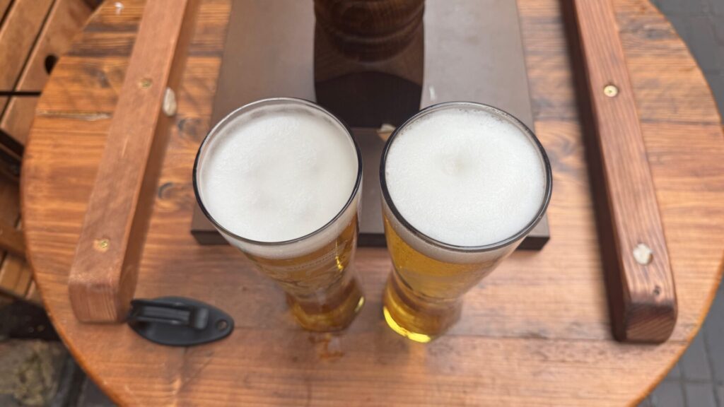 Two freshly poured pints of lager sit side by side on a rustic wooden table at a traditional London pub near Carnaby Street. The beers feature perfectly formed creamy white foam heads that crown the amber-coloured liquid beneath. The wooden table shows natural grain patterns and appears well-worn from years of use, whilst a person's legs in dark trousers are partially visible in the background, creating an authentic pub atmosphere with warm, inviting lighting.