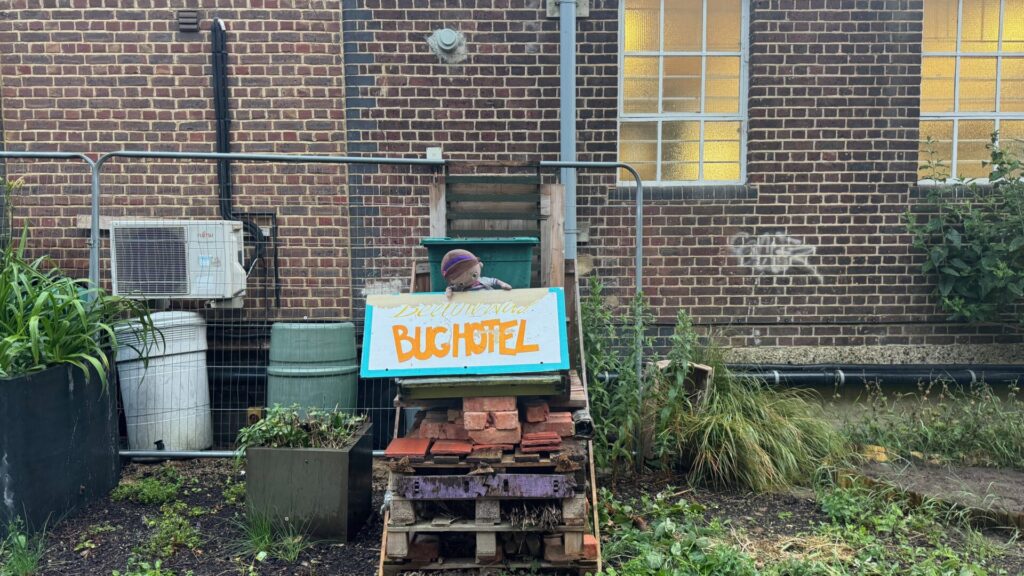 A handmade 'BUGHOTEL' sign in bright orange letters on a turquoise background, marking a bee habitat constructed along The Beeline Way between New Malden and Raynes Park. The insect hotel, designed to provide nesting sites for carpenter and leafcutter bees, sits amongst lush garden vegetation with brick residential buildings visible in the background. A small decorative figure stands guard behind the colourful sign, whilst the surrounding area features typical urban garden elements, including stacked pallets and thriving plant life.