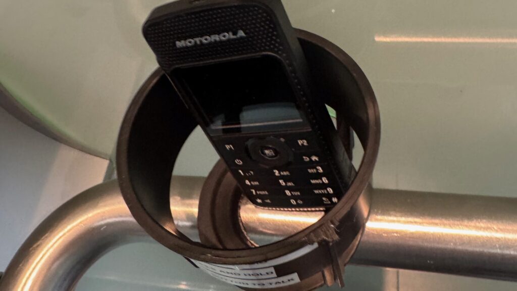 A vintage black Motorola mobile phone with a physical keypad is securely strapped to the handrail inside a lift, serving as an emergency communication device. The phone, featuring the classic button layout and small monochrome display typical of early mobile phones, is mounted on the curved stainless steel handrail against the lift's modern interior. This improvised but practical solution provides a backup communication method for lift emergencies, creating an amusing juxtaposition between old-school mobile technology and contemporary lift design. The phone appears to be properly secured and positioned for easy access by lift users in case of emergency.