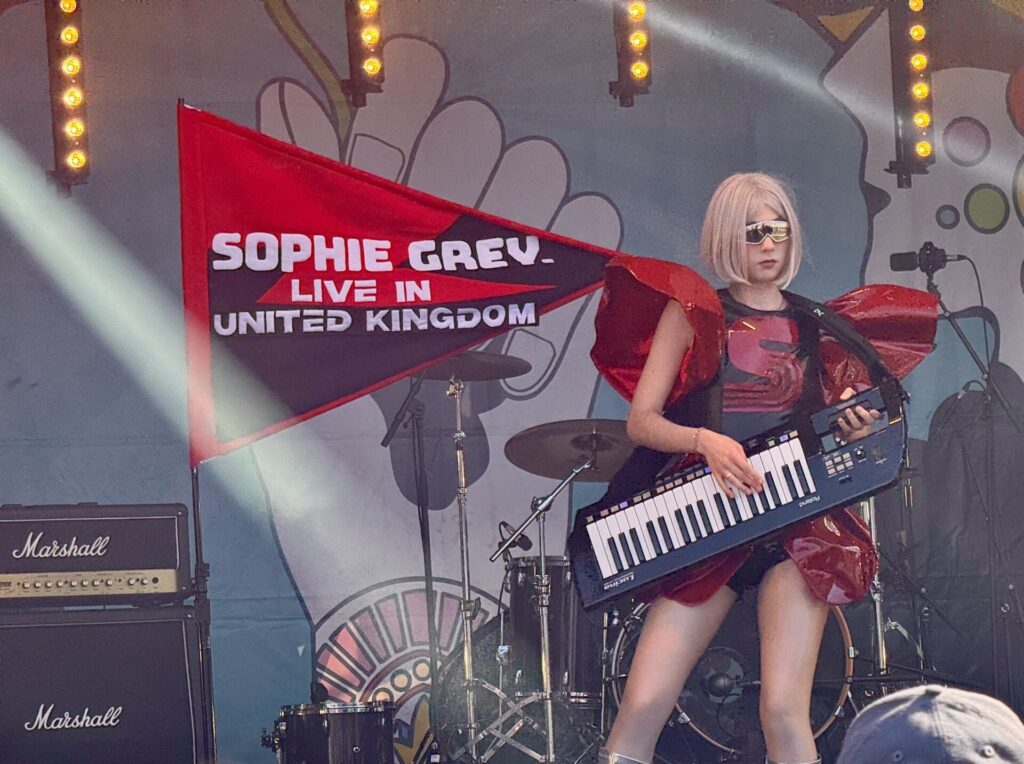 A performer in a red sequined outfit and sunglasses plays a keytar on stage at the 2025 Isle of Wight Festival. A red triangular banner reading "SOPHIE GREY - LIVE IN UNITED KINGDOM" is visible in the background, along with stage lighting, Marshall amplifiers, and drum kit. The artist is captured mid-performance in what appears to be an energetic live music moment at the festival.