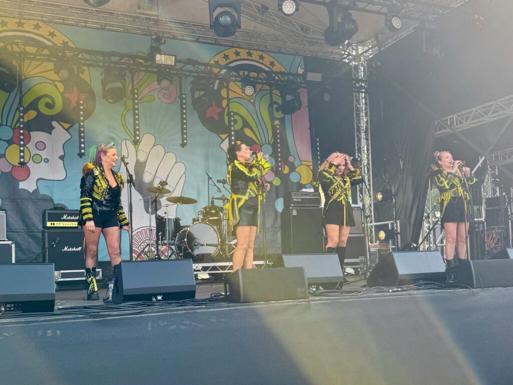 The Queenbees perform on stage at the 2025 Isle of Wight Festival, featuring four female performers in matching black and yellow outfits. The stage backdrop displays a vibrant, psychedelic carnival-style design with swirling patterns, stars, and colourful artwork. The performers are positioned across the stage with microphones, backed by a full drum kit and Marshall amplifiers.