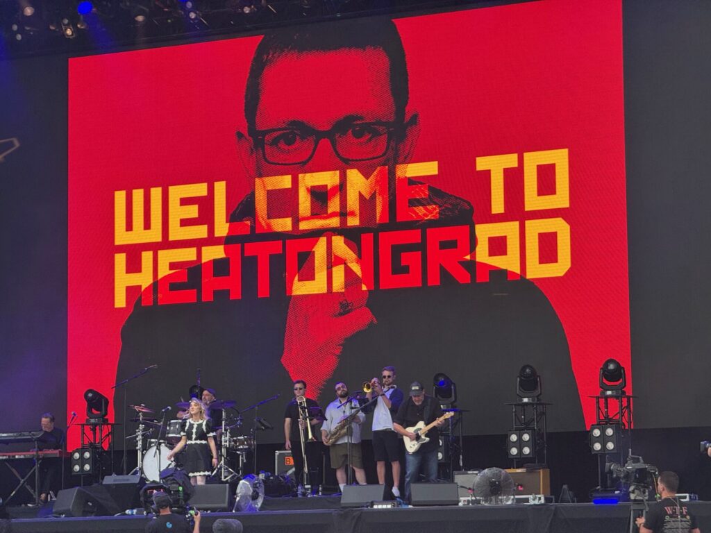 Paul Heaton performs on stage at the 2025 Isle of Wight Festival with his band. The stage backdrop features a bold red design with "WELCOME TO HEATONGRAD" displayed in large yellow letters, alongside a portrait of a man wearing glasses in a halftone print style. The full band setup includes drums, guitars, and brass instruments, with multiple musicians positioned across the stage.