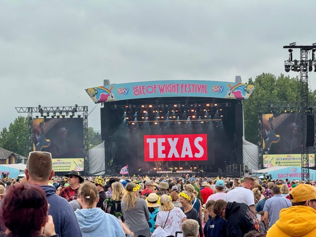 Texas performs on the main stage at the 2025 Isle of Wight Festival, with a large crowd of festival-goers gathered in front of the stage. The stage features the band's name "TEXAS" displayed prominently in white letters on a red background, with the Isle of Wight Festival branding and Sky sponsorship visible on the curved stage canopy above. Large video screens on either side of the stage show close-up footage of the performance, whilst professional lighting rigs illuminate the scene. The diverse audience, wearing a mix of summer clothing and festival attire, fills the field in front of the main stage under an overcast sky