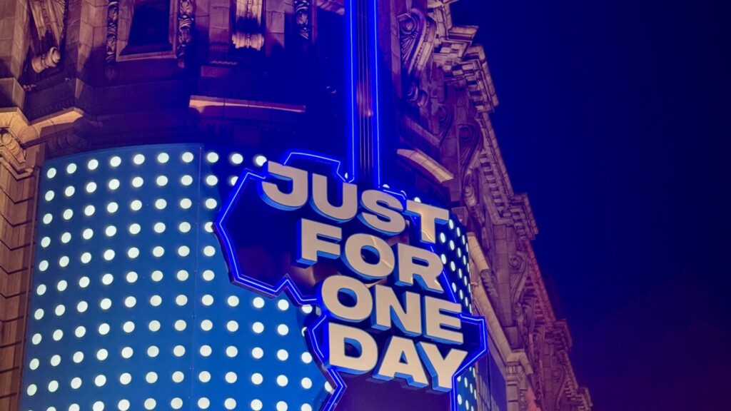 A brightly lit neon sign reading "JUST FOR ONE DAY" in bold white letters with blue neon outlining, mounted on the ornate facade of the Shaftesbury Theatre in London. The sign advertises the musical "Just For One Day" - the story of Live Aid, with decorative lighting elements and the theatre's classical architectural details visible in the background.