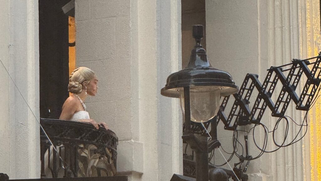 A scene from the revival of "Evita" at the London Palladium shows Rachel Zegler performing as Eva Perón, addressing the crowds from an ornate stone balcony in front of the theatre, to the public watching from below. She is wearing a white strapless gown with her blonde hair styled in a period-appropriate updo, surrounded by classical columns and architecture.