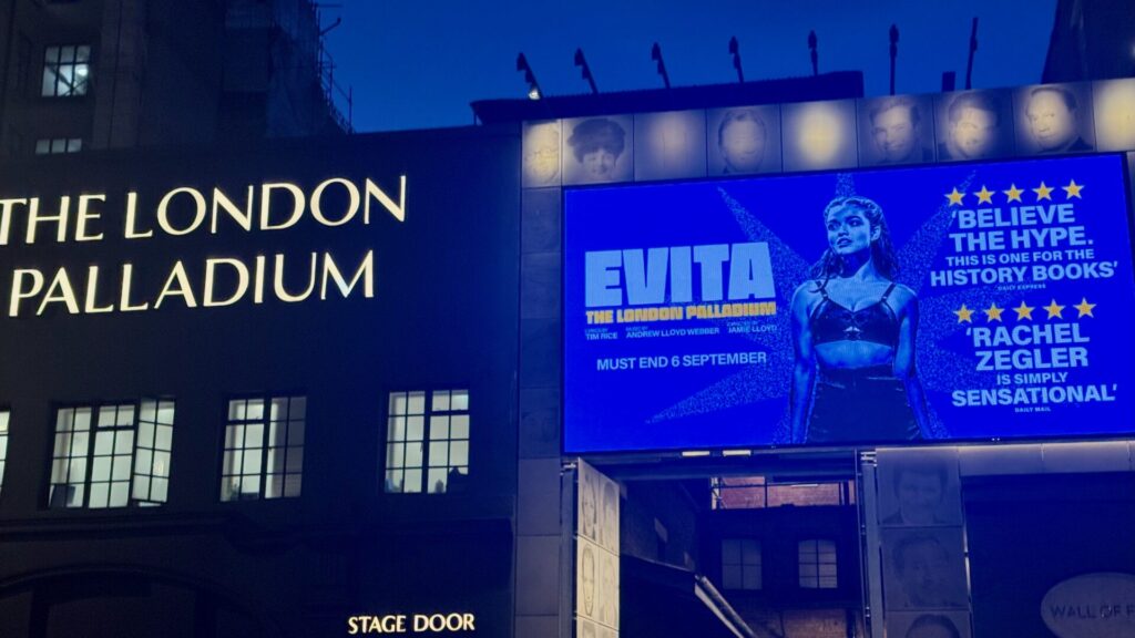 Evening view of the London Palladium's exterior showing the illuminated Evita revival advertisement featuring Rachel Zegler, with the theatre's iconic signage and stage door visible in the atmospheric blue-tinted lighting.