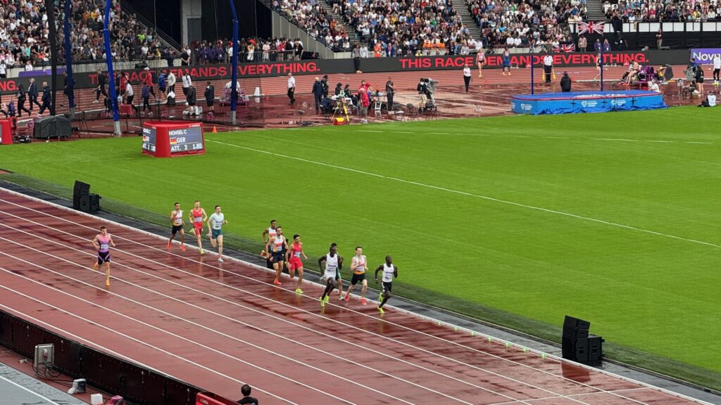 A group of male runners compete in the 800m race at the London Stadium during the Novuna London Athletics Meet. The athletes are running along the curved section of the red running track, with runners spread across multiple lanes wearing various team colours including red, white, and other kit designs. The stadium is packed with spectators in the background stands, and the bright green infield is visible alongside. Electronic timing displays and event branding are positioned around the track perimeter