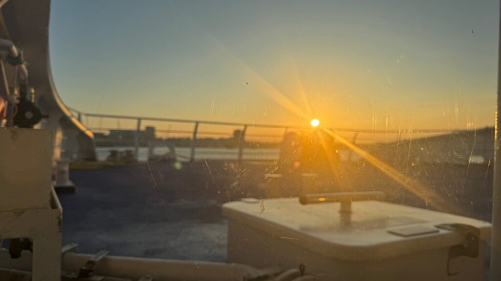 A golden sunset viewed through the salt-stained windscreen of a ferry, with the sun creating dramatic lens flares and highlighting water droplets on the glass. The ferry's white deck furniture and safety railings are visible in the foreground, while the calm waters of the Solent stretch towards the horizon under a gradient sky transitioning from warm orange near the sun to pale blue-grey above.