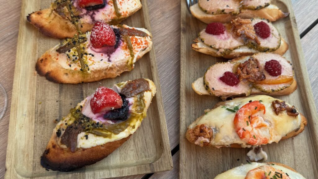 A wooden serving board displaying six gourmet tapas on toasted bread slices, featuring various toppings including sliced meat with strawberries and blackberries, prawns with melted cheese, and other colourful Mediterranean ingredients