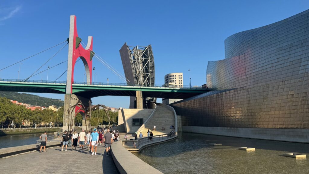 The distinctive titanium-clad exterior of the Guggenheim Museum Bilbao designed by Frank Gehry, with tourists walking along the riverside promenade and the red La Salve Bridge visible in the background