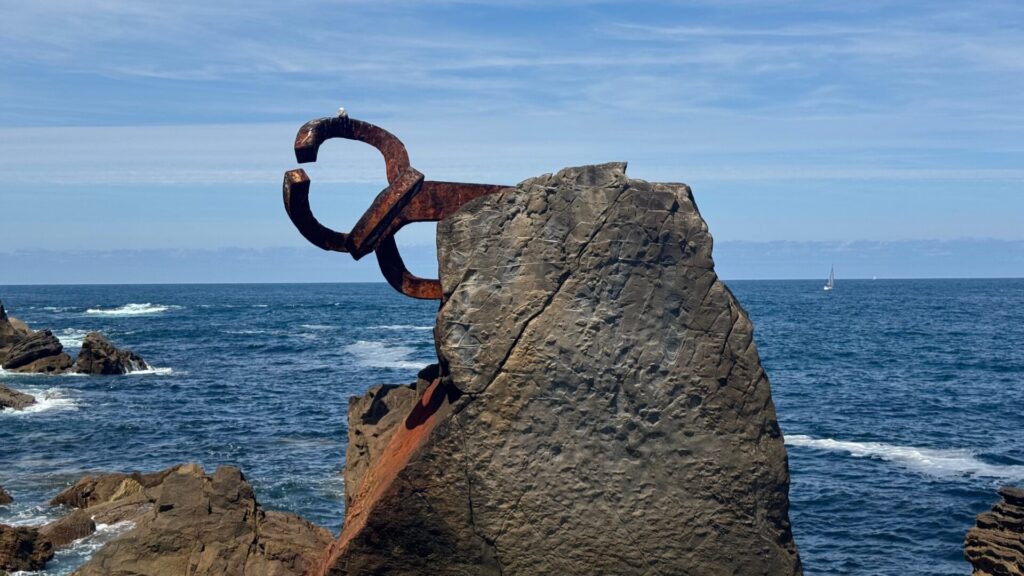 Eduardo Chillida's steel sculpture Peine del Viento installed on rocky coastal cliffs overlooking the Cantabrian Sea in San Sebastián, with waves crashing against the rocks and a small sailing boat visible on the horizon