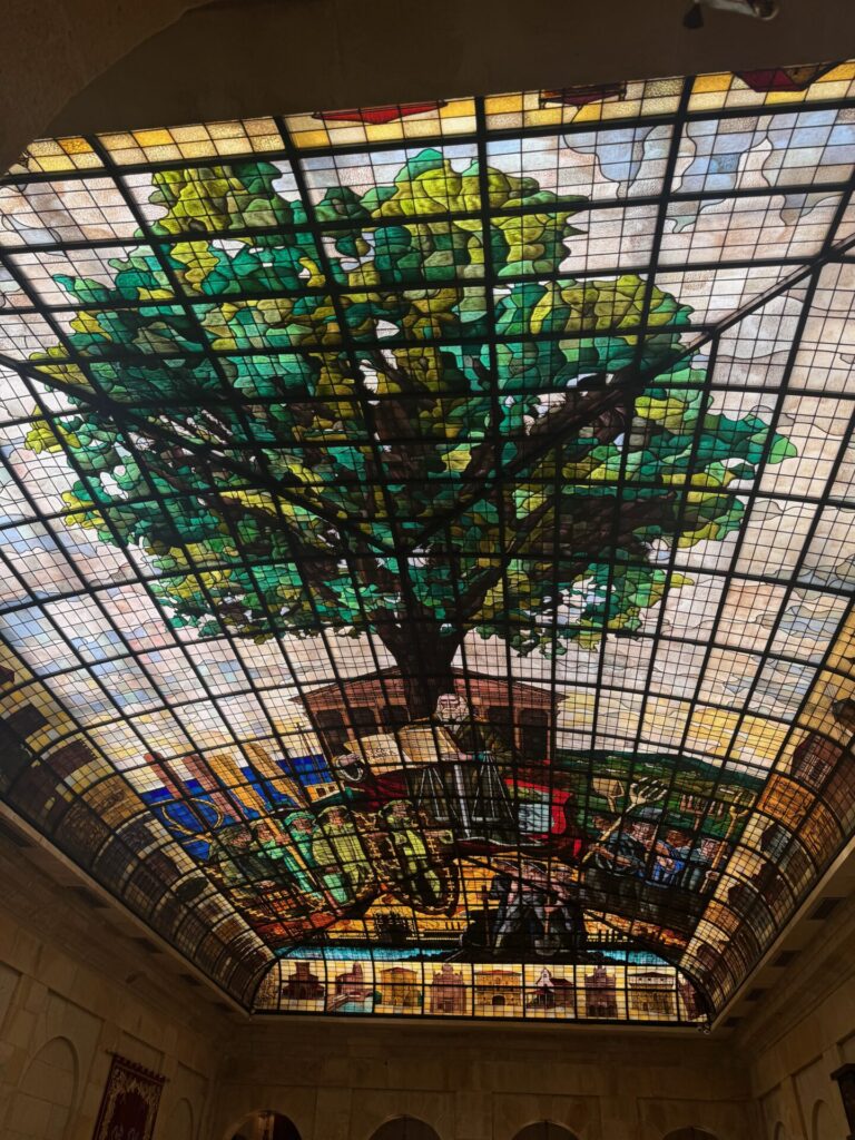 Magnificent stained glass ceiling in the Assembly House of Guernica depicting the sacred Tree of Guernica (Gernikako Arbola) with intricate green and gold foliage against a geometric grid pattern, symbolising Basque freedoms and democratic traditions