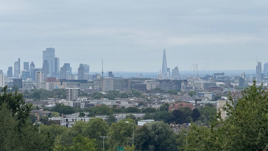 Panoramic view of London's skyline from Hampstead Heath showing the City of London's modern skyscrapers including the Shard and Gherkin rising above a dense canopy of green trees, with residential areas and lower buildings in the foreground under an overcast sky.