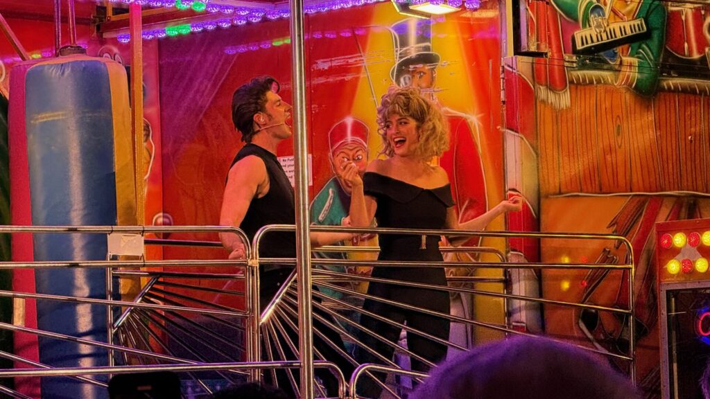 Performers portraying Danny and Sandy stand together at a colourful fairground attraction with vintage carnival decorations and neon lighting