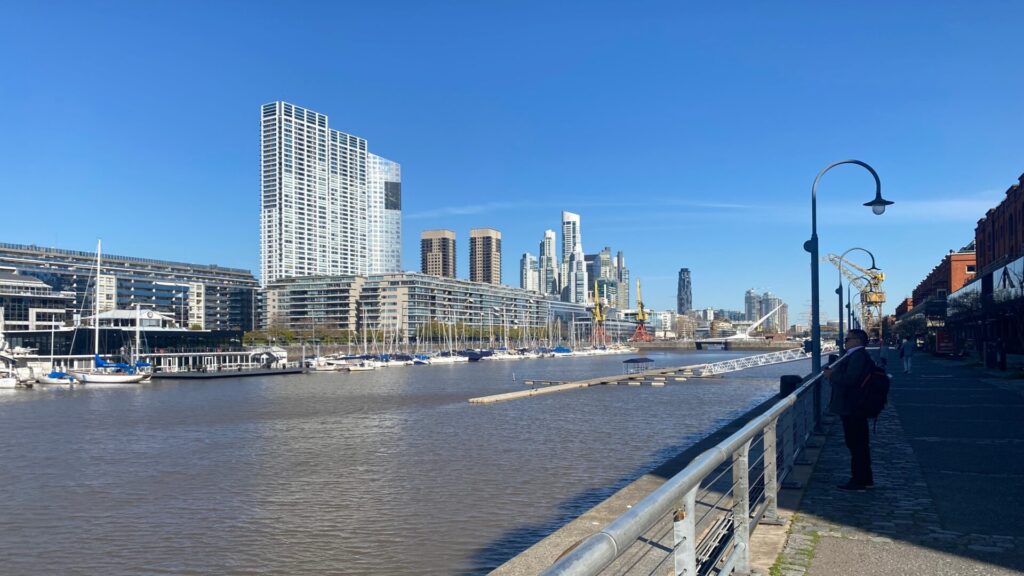 Waterfront view of Puerto Madero, Buenos Aires, showing modern high-rise apartment buildings along the yacht-filled docklands, with a pedestrian promenade in the foreground under clear blue skies