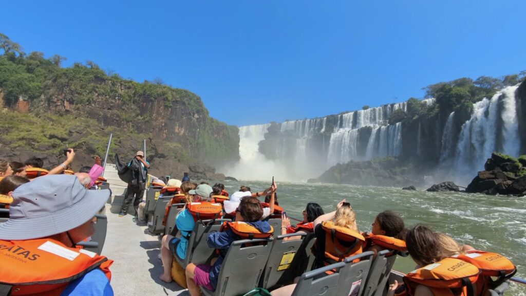 Tour boat full of tourists wearing orange life jackets approaches the base of Iguazu Falls, with massive waterfalls cascading over tiered basalt cliffs surrounded by lush green vegetation and mist rising from the churning water below