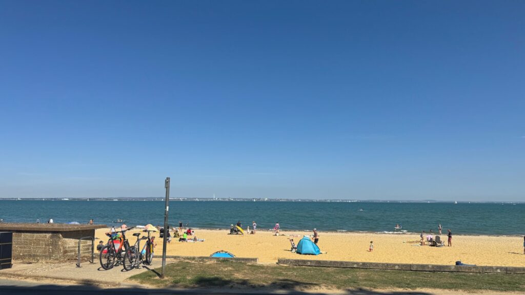 Families and beachgoers relax on Ryde beach on a sunny bank holiday Monday, with colourful windbreaks, beach tents, and people scattered across the golden sand. Bicycles are parked along the promenade in the foreground, whilst the calm blue waters of the Solent stretch towards Portsmouth's distant skyline under clear skies.