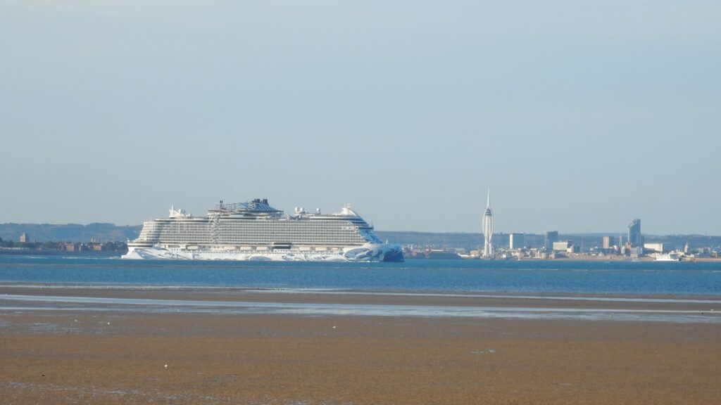 A large cruise ship passes through the Solent waterway with Portsmouth's skyline visible in the background, including the distinctive Spinnaker Tower. The view is captured from Ryde beach at low tide, with wet sand in the foreground and calm blue waters separating the Isle of Wight from the mainland.
