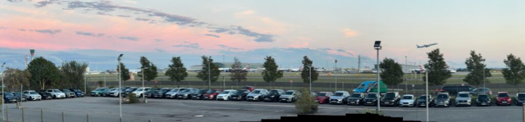 A view across Heathrow Airport's car park at dusk, with rows of parked vehicles in the foreground and aircraft visible on the tarmac beyond the perimeter fence. The sky displays soft pink and blue hues as evening approaches, whilst planes and airport infrastructure create a silhouette against the horizon.