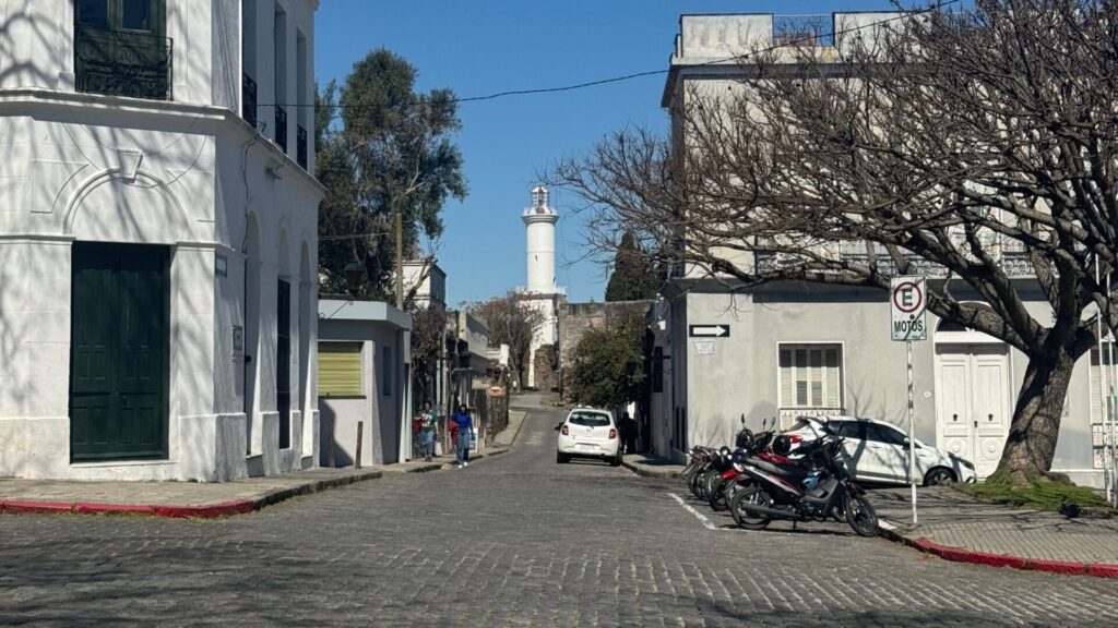 Cobblestone street in Colonia del Sacramento, Uruguay, with colonial-era buildings, a white lighthouse visible in the distance, bare winter trees, and parked motorcycles under clear blue skies