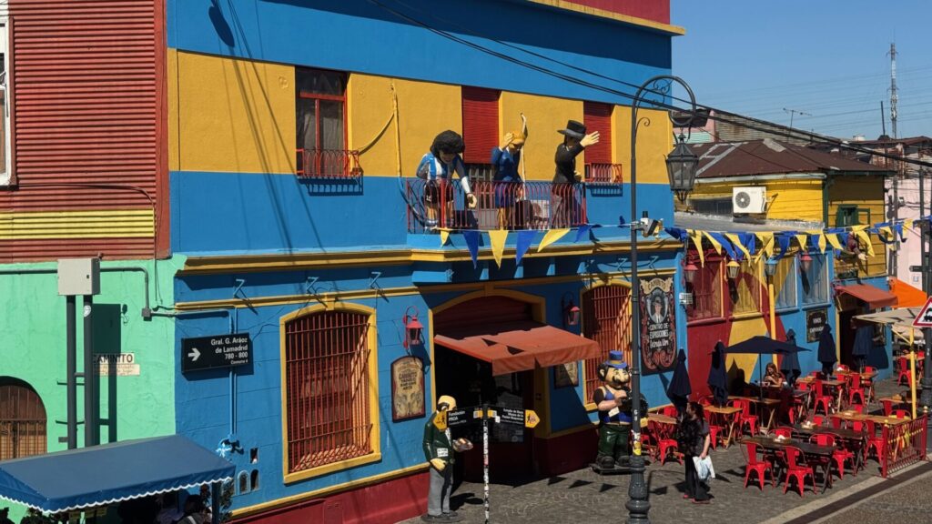 Colourful buildings in El Caminito, Buenos Aires, featuring vibrant blue, yellow, red, and green corrugated metal facades, with life-sized tango dancer figures on balconies, outdoor café seating with red chairs, and tourists exploring the famous street