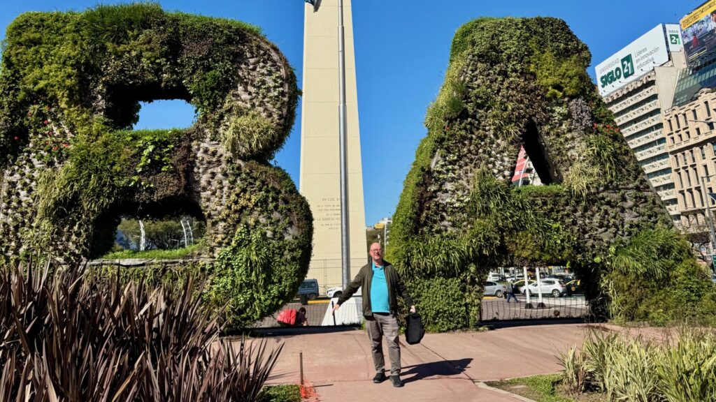 Large living sculpture spelling 'BA' covered in dense green vegetation and plants, with person - the author - standing between letters in Buenos Aires city centre, buildings and blue sky in the background.