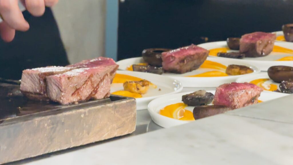 Chef slicing perfectly cooked medium-rare steak on a wooden board at Fogón Asado's chef's counter in Buenos Aires, with plated portions of grilled meat and vibrant yellow sauce arranged on white plates in the foreground