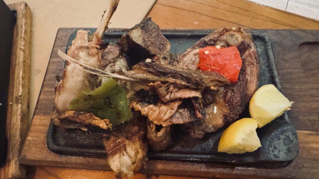Grilled Patagonian lamb served on a cast iron griddle with bone attached, accompanied by roasted red pepper, grilled vegetables, and lemon wedges, presented on a rustic wooden board in El Calafate, Argentina