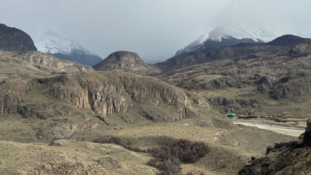 Rugged Patagonian landscape near El Chaltén showing rocky outcrops and golden grassland in the foreground, with snow-capped mountain peaks visible in the distance under an overcast sky, and a small green-roofed building in the valley