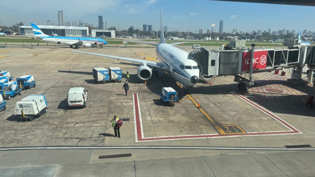 Aerolíneas Argentinas aircraft parked at gate with jet bridge attached at Aeroparque Jorge Newbery, with ground crew and service vehicles on the tarmac, Buenos Aires city skyline visible in the background under clear skies