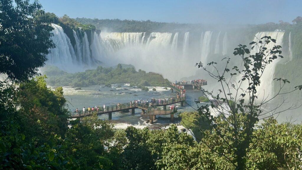Panoramic view of Iguaçu Falls with multiple cascading waterfalls creating massive spray, viewing platforms filled with tourists extending into the river surrounded by lush green subtropical forest under clear blue skies