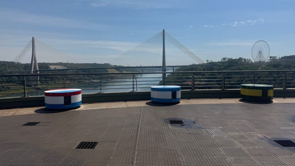 Viewing platform at Hito Tres Fronteras with three cylindrical monuments painted in the colours of Argentina, Paraguay, and Brazil, overlooking the confluence of rivers with a cable-stayed bridge and observation wheel visible in the distance under blue skies