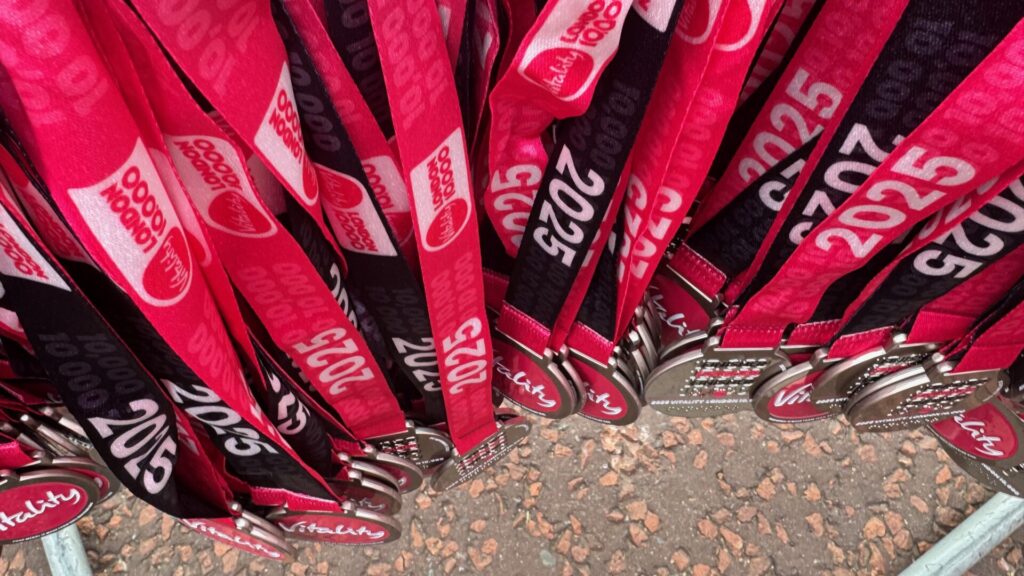 Collection of Vitality 10,000 finisher medals with red and black lanyards displaying '2025' and event branding, arranged in a radiating pattern on the fence awaiting distribution to race participants