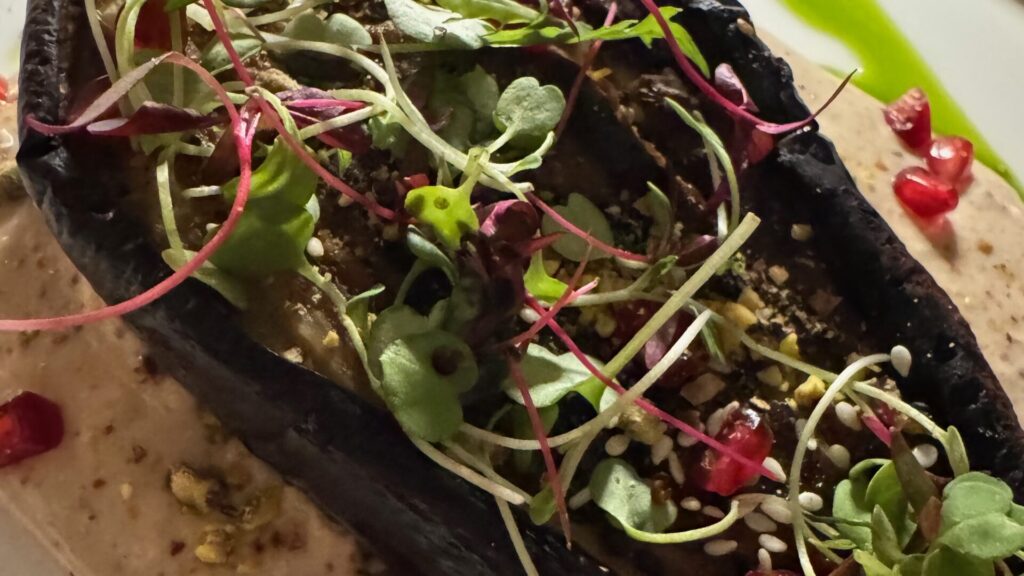 A close-up photograph of a charred aubergine dish topped with colourful microgreens in shades of green and pink, sesame seeds, and dots of red sauce, served on a pale ceramic plate with decorative sauce dots around the edges.
