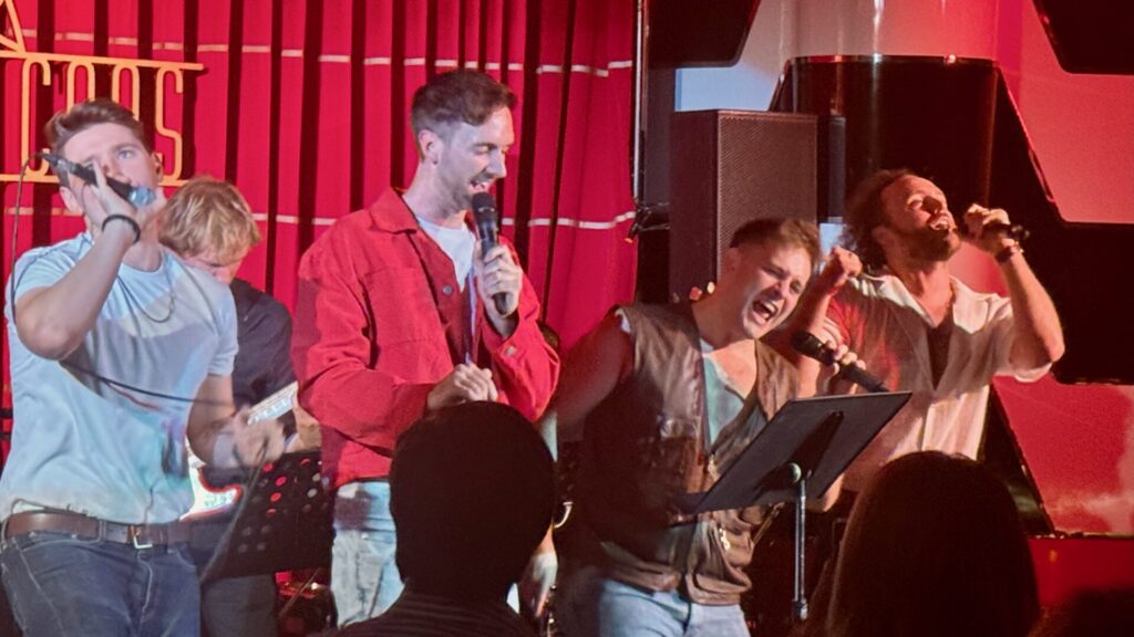 Five male performers singing enthusiastically into microphones on stage at The Crazy Coqs, with a red curtain backdrop and neon signage, performing songs from Bruce Springsteen's catalogue