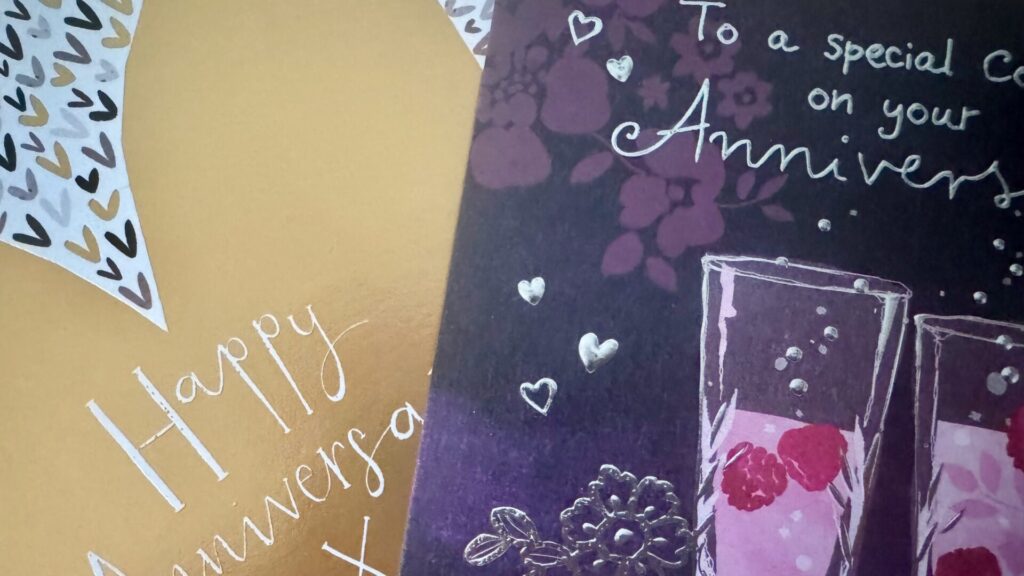 A collage showing portions of two anniversary cards side by side. The left card has a mustard yellow background with white text reading "Happy Anniversary" and a decorative heart pattern border. The right card has a dark purple background with pink flowers, white hearts, text reading "To a special couple on your Anniversary", and an illustration of two champagne glasses with raspberries and bubbles.