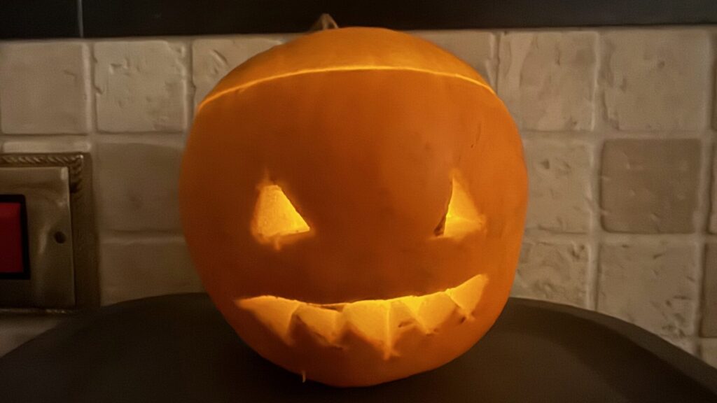 A carved Halloween pumpkin with triangular eyes and a jagged, toothy grin, illuminated from within by a warm glow, sitting on a dark surface against a white brick wall.