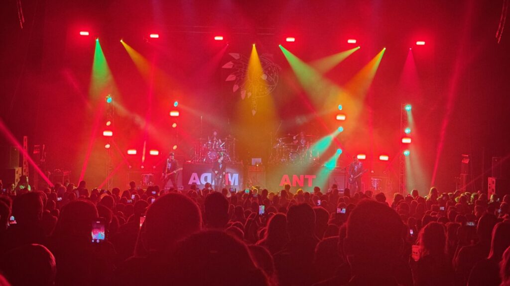 A concert photograph showing a large crowd watching a performance at the Hammersmith Apollo. The stage is bathed in dramatic red and orange lighting with beams cutting through atmospheric haze. Multiple spotlights create geometric patterns across the venue. The band's name "ADAM ANT" is visible on illuminated panels on stage, with silhouettes of performers and their equipment visible through the colourful lighting. Audience members in the foreground are holding up phones to capture the moment.