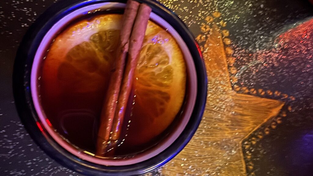 A purple-rimmed mug of mulled wine with two cinnamon sticks and an orange slice, photographed from above on a wooden table with colourful reflections