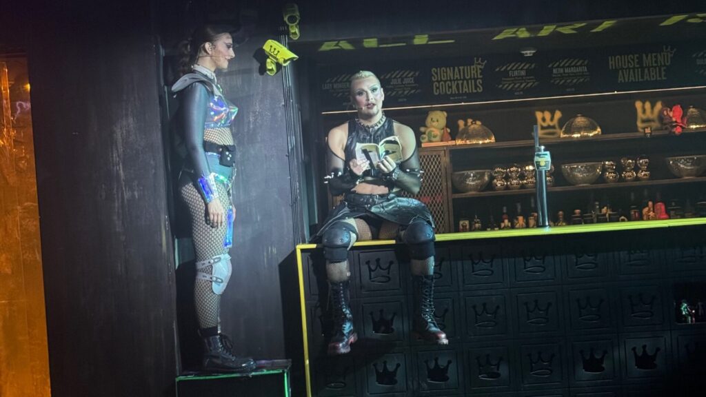 Two performers in futuristic cyberpunk costumes at a dystopian bar set. One performer stands on an elevated platform wearing fishnet stockings, metallic blue accessories, and knee pads, whilst the other sits on the illuminated bar counter in similar edgy attire with protective gear. Behind them is a dark bar backdrop with neon yellow signage advertising signature cocktails, shelves displaying quirky figurines and glassware. The aesthetic combines industrial grunge with neon lighting in green and yellow tones.