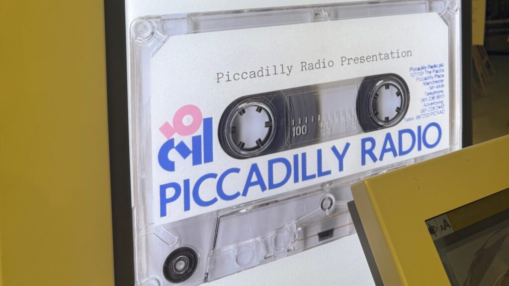 Clear cassette tape labeled 'Piccadilly Radio Presentation' showing analogue tape reels and counter markings, displayed at exhibition with yellow background