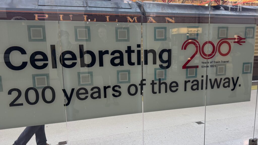 A close-up of a frosted window platform divider at Victoria Station. Bold black text reads "Celebrating 200 years of the railway." To the right, a stylised red logo displays the number 200, with the British Rail double-arrow symbol integrated into the design. The words "Pullman" are visible on the dark coach work above the window, and people are faintly reflected in the glass